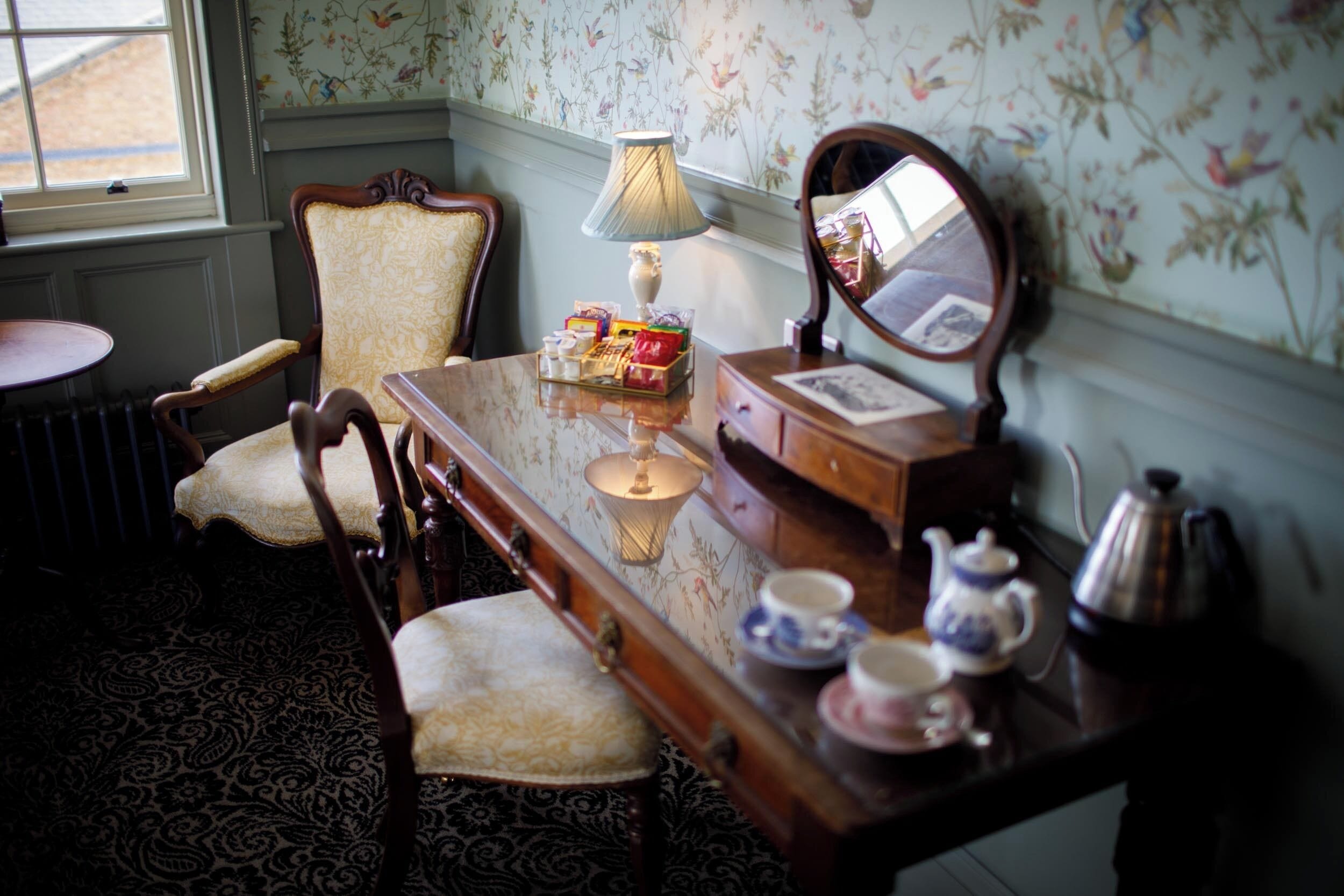 A room with a wooden desk, two chairs, a lamp, a mirror, and a radiator.