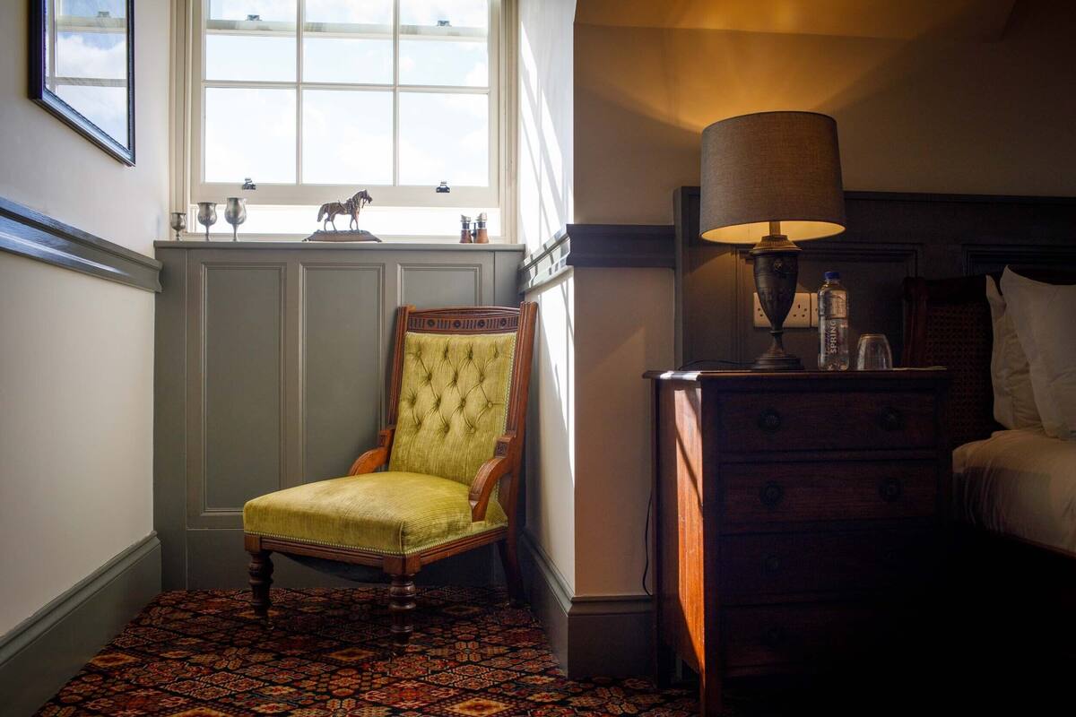 A room with a green upholstered chair, a wooden nightstand with a lamp, and a bed with white bedding.
