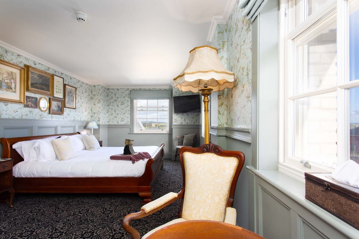 A hotel room with a large bed, a chair, a desk, a lamp, a TV, and a window.