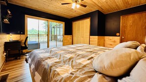 Um quarto com teto de madeira, uma cama com colcha, uma escrivaninha com cadeira e uma porta de vidro deslizante para uma área externa.