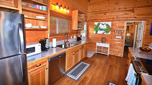 Une cuisine rustique aménagée dans une cabane en bois, avec un réfrigérateur, un four à micro-ondes et un lave-vaisselle en acier inoxydable, des placards en bois et une fenêtre équipée de stores.