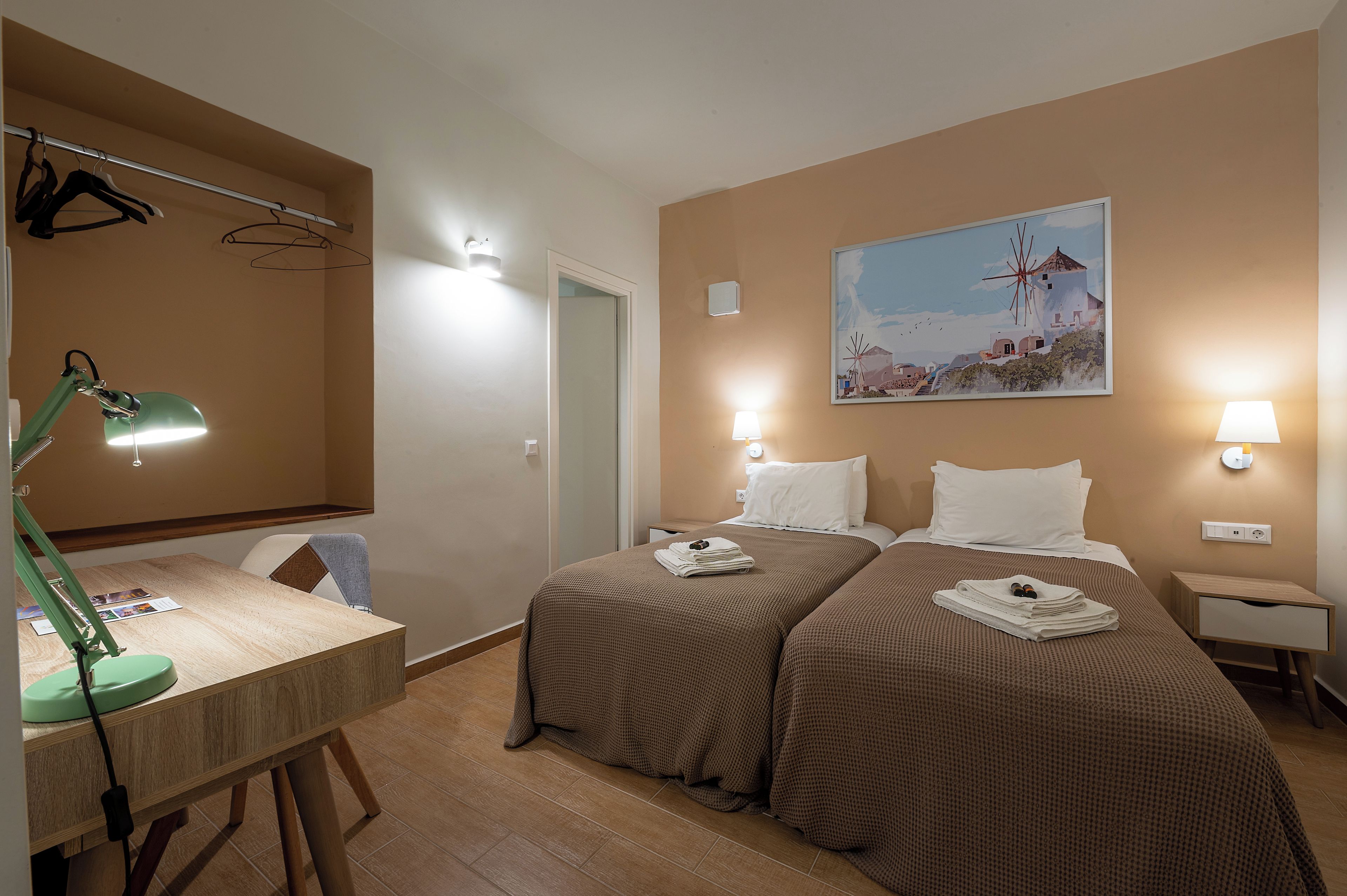 A hotel room with two beds, a desk with a lamp, a painting of a windmill, and wall-mounted lights.