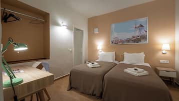 A hotel room with two beds, a desk with a lamp, a painting of a windmill, and wall-mounted lights.