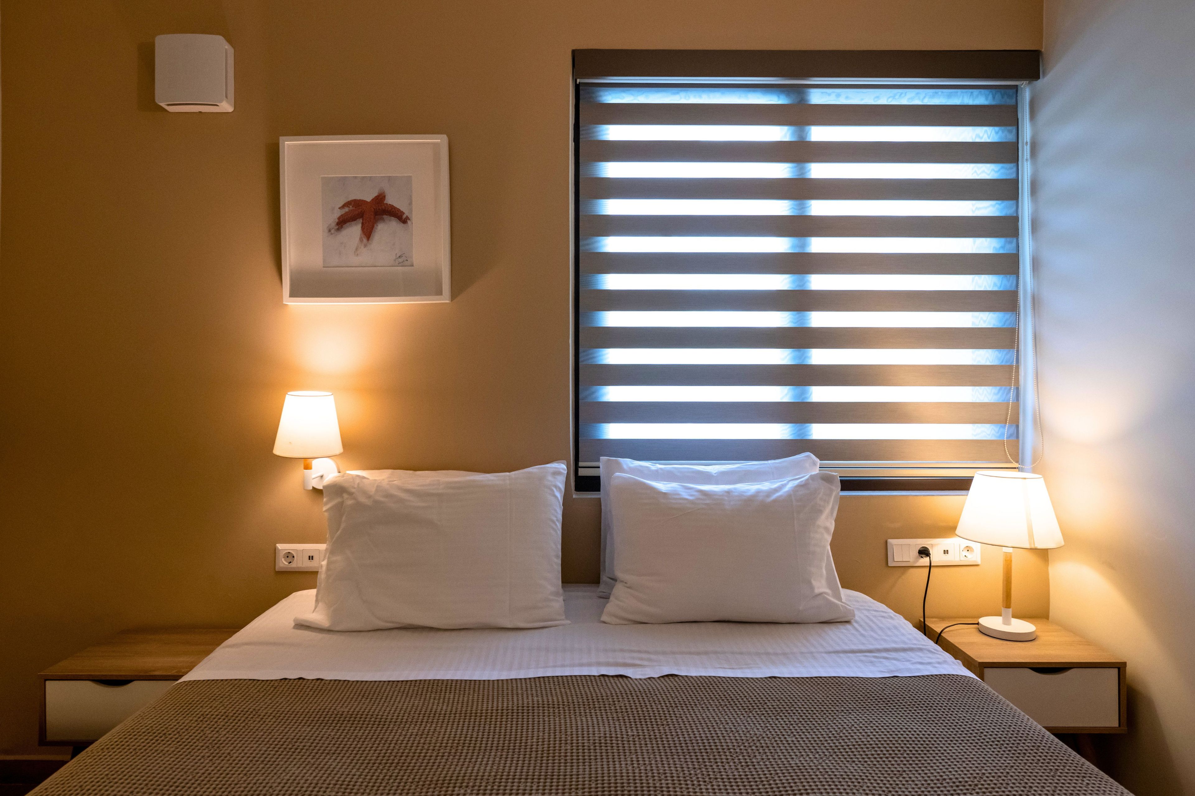 Ein Hotelzimmer mit einem Bett, zwei Nachttischlampen, einem Fenster mit Jalousien, einer Wandleuchte und einem gerahmten Bild.