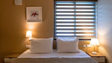 A hotel room with a bed, two bedside lamps, a window with blinds, a wall-mounted light, and a framed picture.