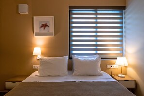 A hotel room with a bed, two bedside lamps, a window with blinds, a wall-mounted light, and a framed picture.