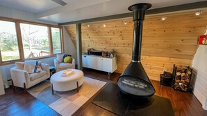 A cozy living room with a wood-burning stove, a sofa, a coffee table, and a view of the outdoors through large windows. - Modern A-Frame on fenced, wooded Lot. Pet friendly! (East Tawas)