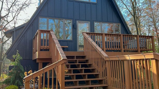 A wooden cabin with a steeply pitched roof, a central chimney, and a front porch with wooden steps leading to a deck. - Modern A-Frame on fenced, wooded Lot. Pet friendly! (East Tawas)