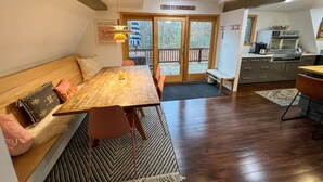 A cozy dining area with a wooden table and bench, a kitchen with modern appliances, and a view through the glass doors. - Modern A-Frame on fenced, wooded Lot. Pet friendly! (East Tawas)
