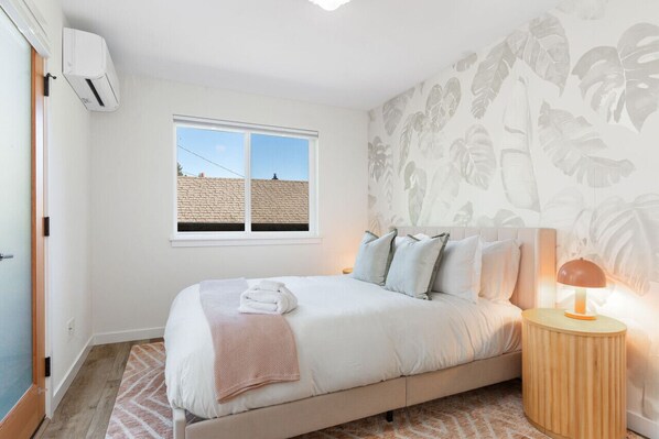 Une chambre à coucher avec un lit, une table de chevet avec une lampe, une fenêtre et un mur orné d’un motif de feuilles.