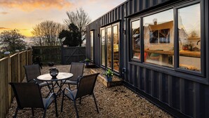 A modern outdoor patio with a round table and chairs, a wooden fence, and a view of a house through large windows. - 1 bedroom accommodation in East Ilsley (East Ilsley)