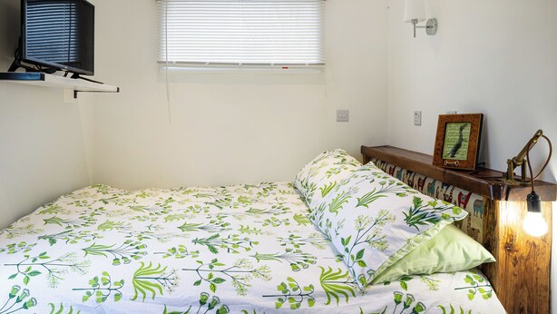 A bedroom with a bed, a television mounted on the wall, a window with blinds, a bedside lamp, and a small shelf with a picture frame. - 1 bedroom accommodation in East Ilsley (East Ilsley)