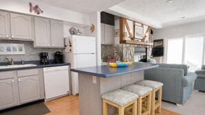 A modern kitchen with a central island, a dining area with a blue sofa, and a stone fireplace. - Ocean Reef 4-B2, Oceanfront Condo in Emerald Isle, NC! (Emerald Isle)
