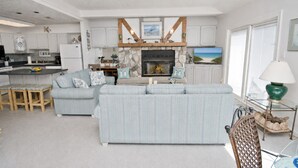 A modern kitchen with white cabinets, a dining area with a light blue sofa, and a living room with a large sofa and a fireplace. - Ocean Reef 4-B2, Oceanfront Condo in Emerald Isle, NC! (Emerald Isle)