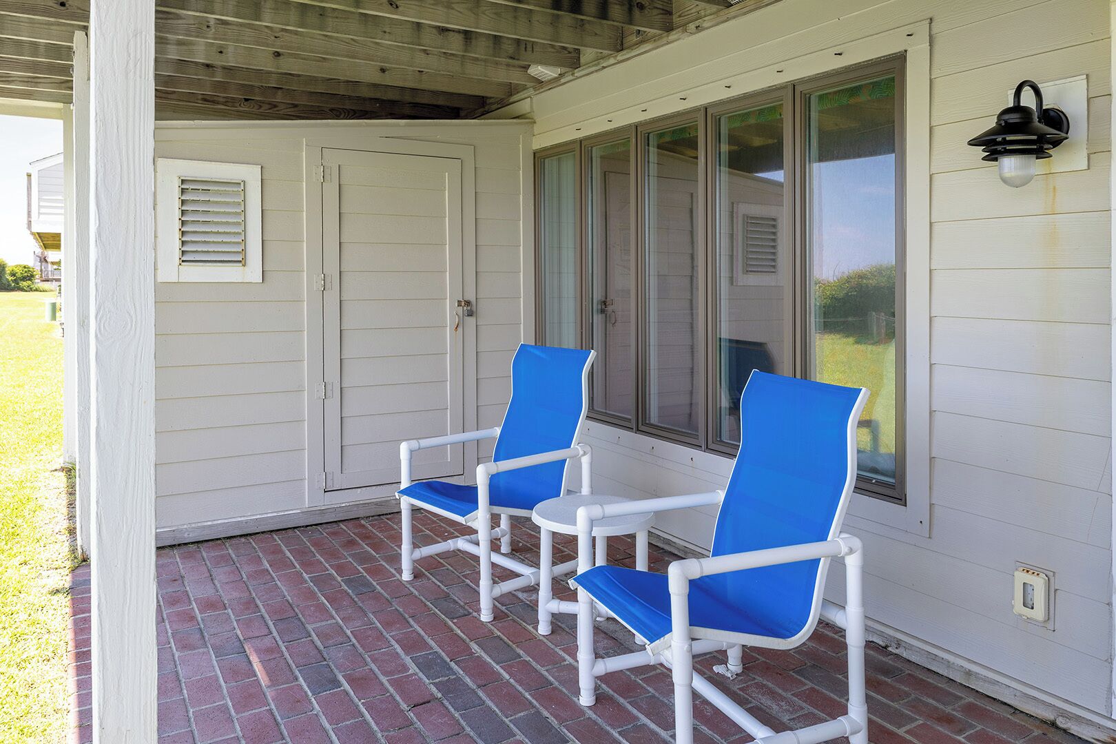 A covered porch with two blue chairs and a small table, white siding, and a window with blinds.