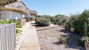 A modern building with a wooden deck and a pathway leading to a natural area with shrubs and plants. - Oceans #12, Oceanside Condo in Pine Knoll Shores, NC! (Pine Knoll Shores)
