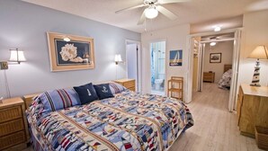 A bedroom with a large bed, a ceiling fan, and a door leading to a bathroom. - Oceans #12, Oceanside Condo in Pine Knoll Shores, NC! (Pine Knoll Shores)