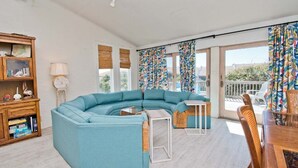 A spacious living area with a teal sectional sofa, a wooden coffee table, and a dining table with chairs. There is a wooden cabinet with decorative items and a lamp. The room has large windows with colorful curtains and a view of the outdoors. - Oceans #12, Oceanside Condo in Pine Knoll Shores, NC! (Pine Knoll Shores)
