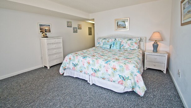 Une chambre à coucher avec une couvre-lit à motifs floraux, une commode blanche, une table de chevet avec une lampe et des tableaux encadrés accrochés aux murs.