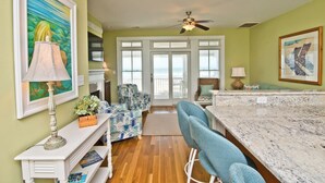 A room with a wooden floor, a ceiling fan, a white fireplace, a bar area with a marble countertop, and a view through large windows. - Palm Cottage I At The Grove, Oceanfront House in Atlantic Beach, NC! (Atlantic Beach)