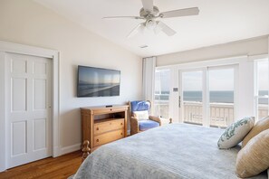 A bedroom with a bed, a TV, a wooden dresser, and a balcony with a view of the ocean.