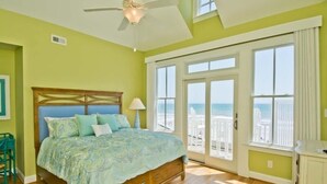 Une chambre avec un lit, un ventilateur de plafond, une vue sur la plage grâce à de grandes fenêtres et un plancher en bois.