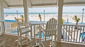 Un espace extérieur abrité, doté de chaises Adirondack blanches et d’une table, qui offre une vue sur une plage avec une piscine et des parasols.