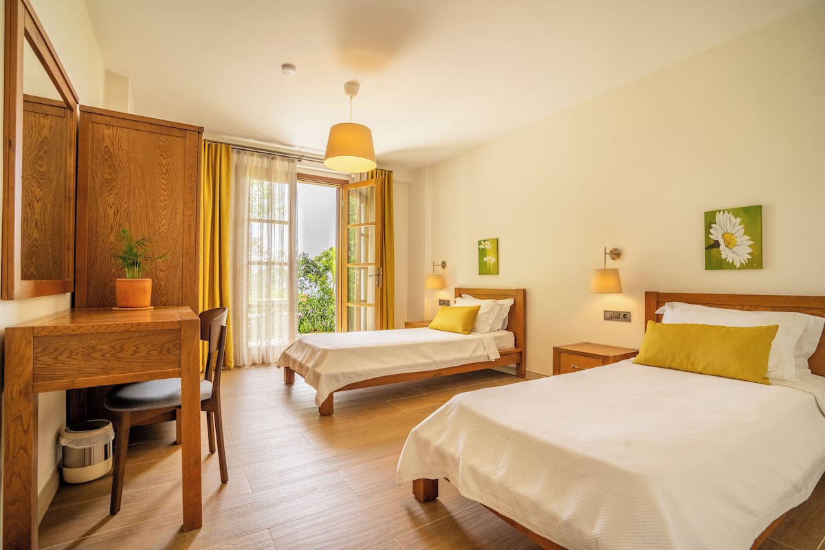 A hotel room with two beds, a desk, a chair, a potted plant, and a view of greenery outside.