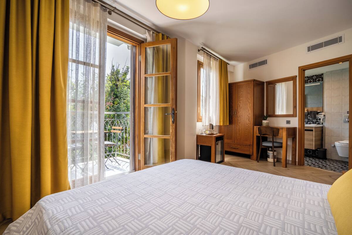 A bedroom with a large bed, a wooden desk, a mirror, and a view of the outdoors.