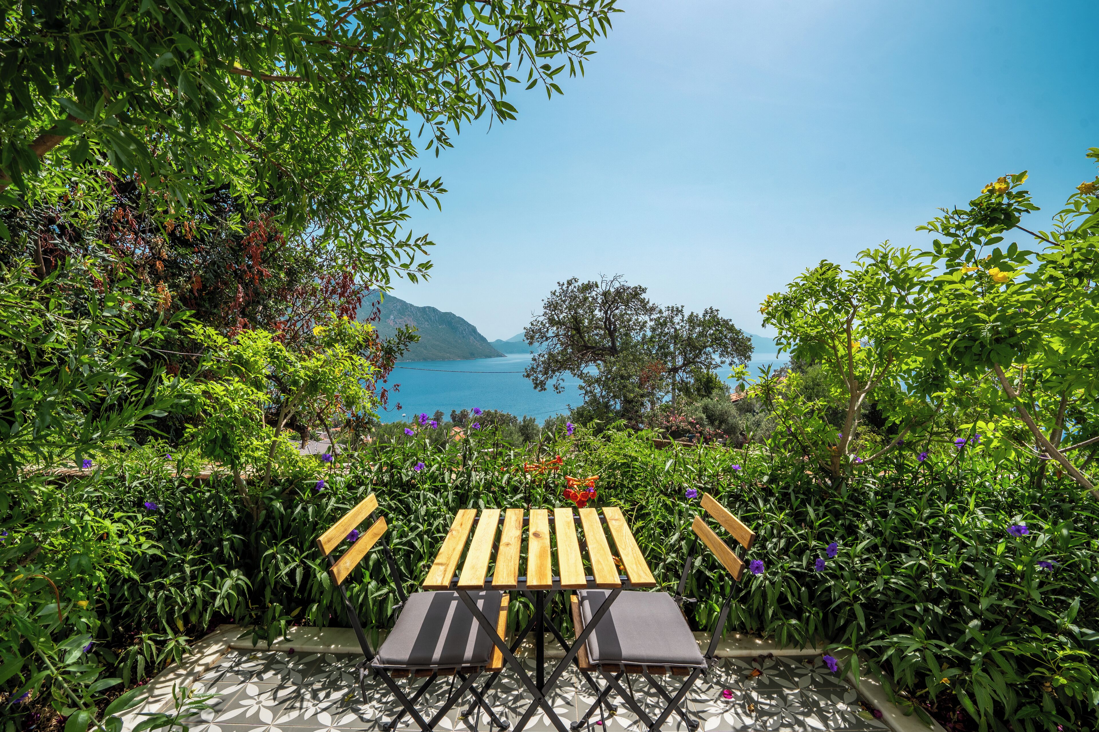 A wooden table and chairs set in a garden with a view of the sea and mountains.