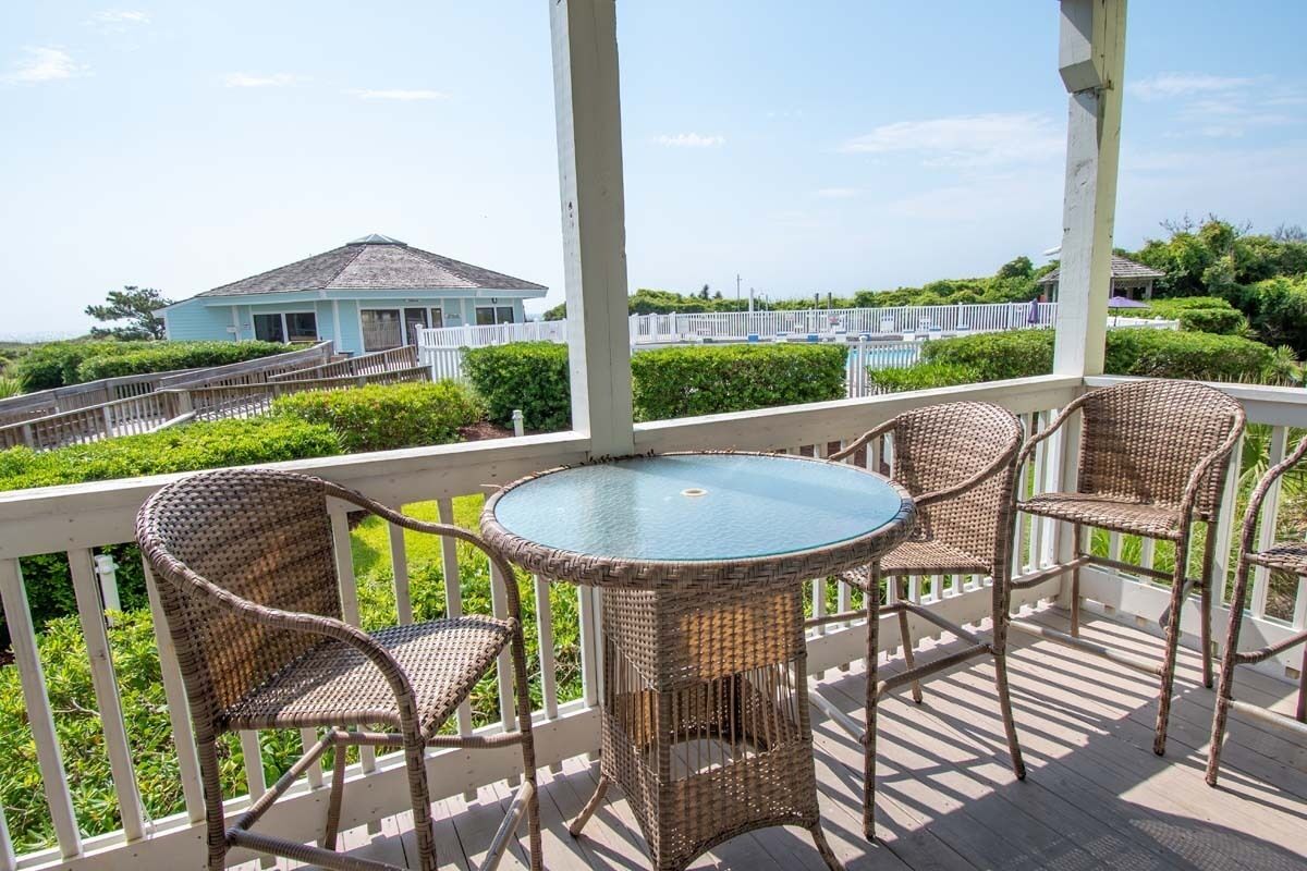 A covered outdoor area with wicker furniture and a glass table, overlooking a house and a fenced area with greenery.