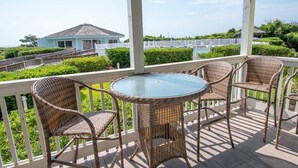 A covered outdoor area with wicker furniture and a glass table, overlooking a house and a fenced area with greenery. - Pebble Beach A-107, Oceanfront Condo in Emerald Isle, NC! (Emerald Isle)