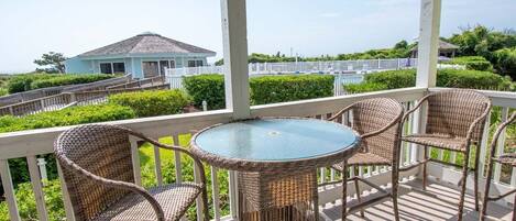 A covered outdoor area with wicker furniture and a glass table, overlooking a house and a fenced area with greenery.
