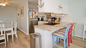 A modern kitchen with white cabinetry, a stainless steel refrigerator, a granite countertop, and a small island with colorful bar stools. - Pebble Beach A-107, Oceanfront Condo in Emerald Isle, NC! (Emerald Isle)