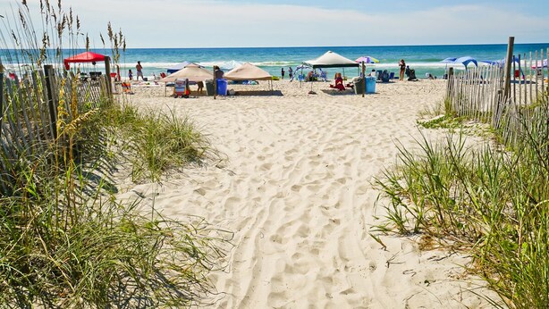 A sandy beach with beach umbrellas and chairs, a wooden fence, and the ocean in the background. - Pebble Beach A-107, Oceanfront Condo in Emerald Isle, NC! (Emerald Isle)