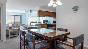 A dining area with a wooden table and chairs, a ceiling fan, and a kitchen in the background. - Pebble Beach A-204, Oceanfront Condo in Emerald Isle, NC! (Emerald Isle)