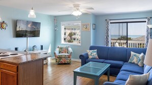 A living room with a blue sofa, a coffee table, a TV, and a kitchen area with a stove. - Pebble Beach A-207, Oceanfront Condo in Emerald Isle, NC! (Emerald Isle)