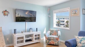 A living room with a flat-screen TV, a white entertainment unit, a floral armchair, a blue sofa, and a window with a view of a building. - Pebble Beach A-207, Oceanfront Condo in Emerald Isle, NC! (Emerald Isle)