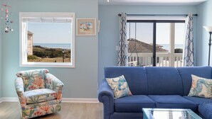 A living room with a view of the beach, a blue sofa, a floral armchair, and a ceiling fan. - Pebble Beach A-207, Oceanfront Condo in Emerald Isle, NC! (Emerald Isle)
