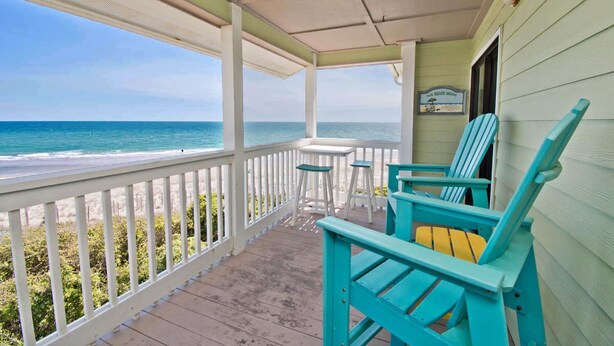 Eine überdachte Veranda mit türkisfarbenen Adirondack-Stühlen und einem kleinen Tisch mit Blick auf einen Strand.
