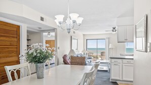 A modern kitchen with white cabinets, a dining area with a white table and chairs, and a view of the ocean through large windows. - Pebble Beach A-304, Oceanfront Condo in Emerald Isle, NC! (Emerald Isle)