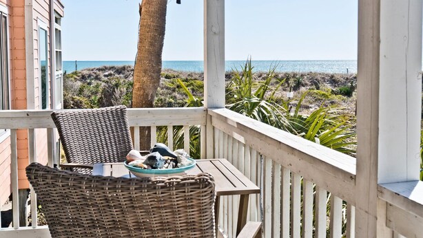 A wooden deck with wicker furniture and a table, overlooking a beach with a palm tree and vegetation.