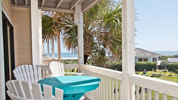 A covered outdoor area with white chairs and a turquoise table, overlooking a beach and a house. - Pebble Beach D-202, Oceanfront Condo in Emerald Isle, NC! (Emerald Isle)