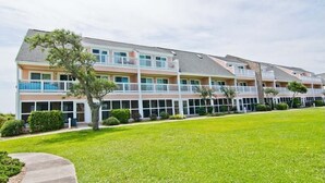 A multi-story hotel with balconies, a well-maintained lawn, and a clear sky. - Pebble Beach 1-110, Oceanside Condo in Emerald Isle, NC! (Emerald Isle)