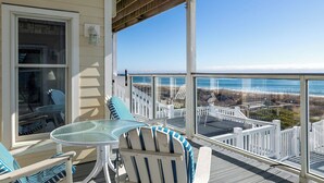 A covered outdoor area with a glass table and chairs, a view of the ocean, and a white railing. - Pelican Perch, Oceanfront House in Emerald Isle, NC! (Emerald Isle)