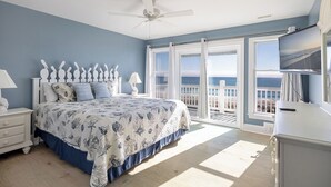 A bedroom with a bed, a nightstand, a dresser, a ceiling fan, and a large window with a view of the beach. - Pelican Perch, Oceanfront House in Emerald Isle, NC! (Emerald Isle)