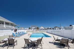 A pool area with lounge chairs and a table, a fenced deck, and a building in the background.