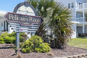 Une enseigne indiquant « Pelican’s Roost Condos », avec une vue sur un immeuble et sur les aménagements paysagers.