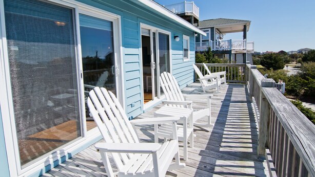 A seaside house with a wooden deck, white chairs, and a balcony overlooking the ocean.