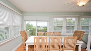 A dining area with a white table and wicker chairs, large windows with blinds, and a ceiling fan.